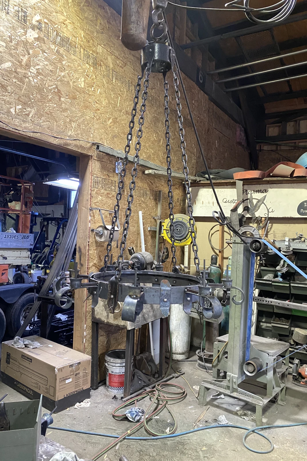 A large, custom-hand-forged black wrought iron chandelier in a medieval style, featuring a heavy circular band with decorative scrollwork and hanging from thick industrial chains in the metalworking shop.
