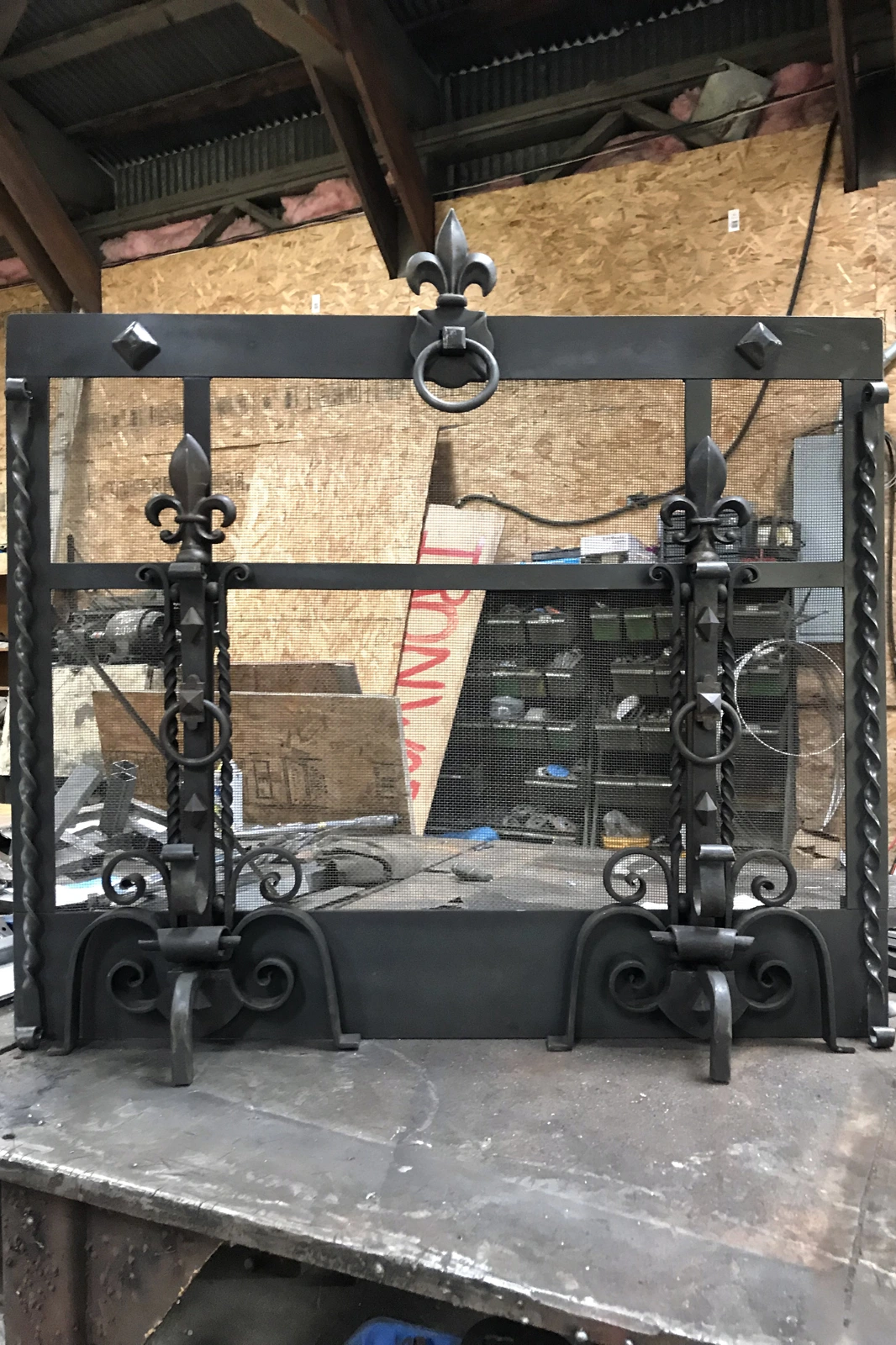 A custom hand-forged black wrought iron fireplace screen featuring intricate fleur-de-lis finials, twisted iron borders, and decorative scrollwork feet. | Chip and Dale's Iron Works