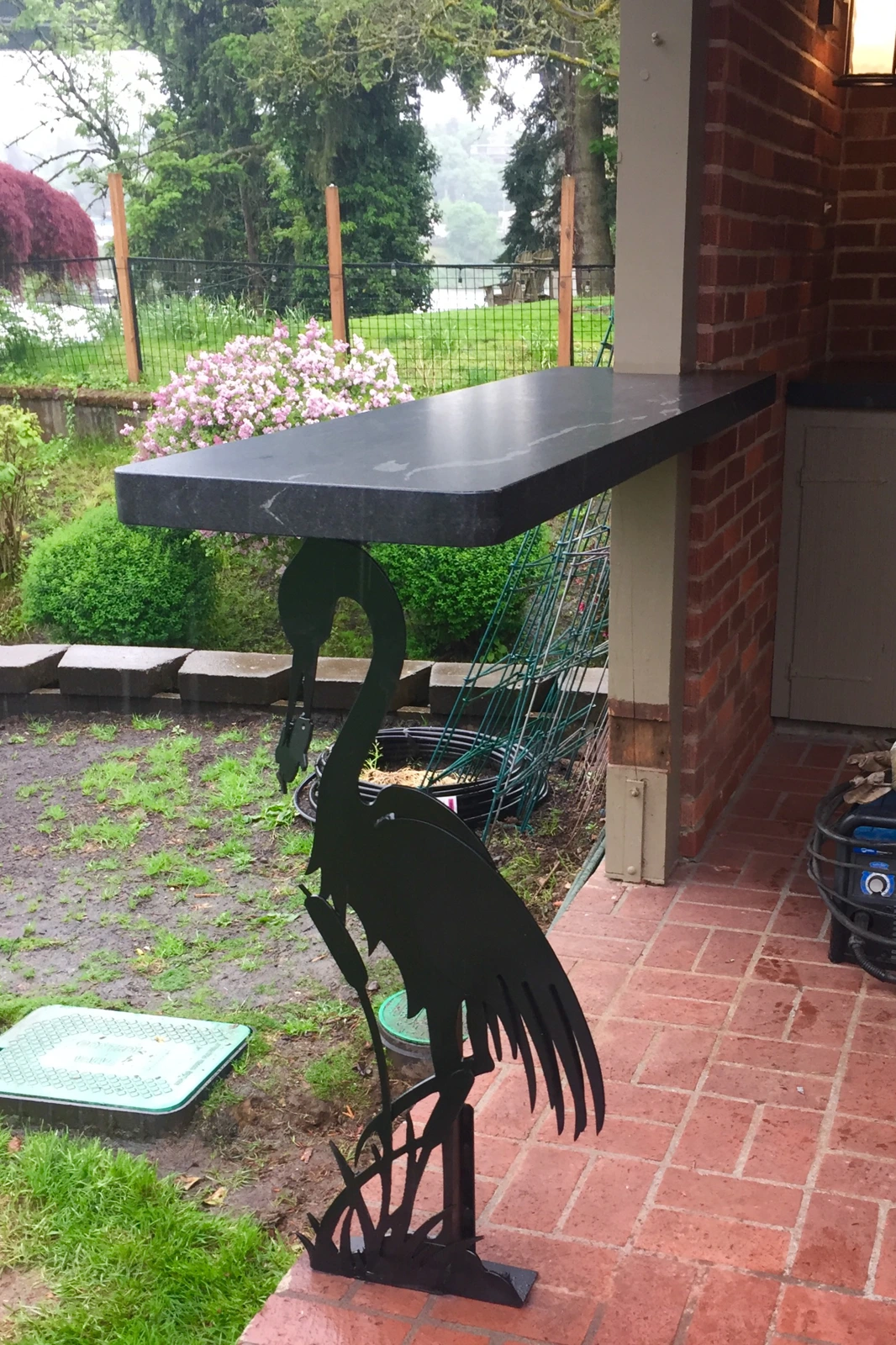 A custom-handcrafted black metal bar support in the silhouette of a blue heron with cattails, holding up a black granite outdoor countertop on a red brick patio.
