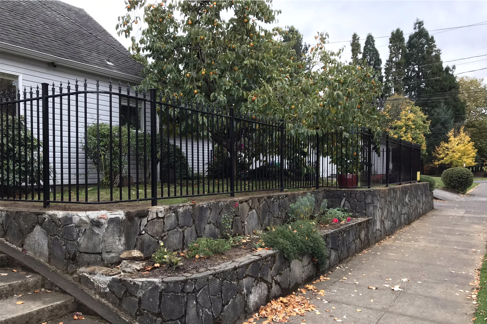 A black ornamental iron fence with spear-top finials installed on top of a multi-tiered stone retaining wall bordering a residential garden and sidewalk.