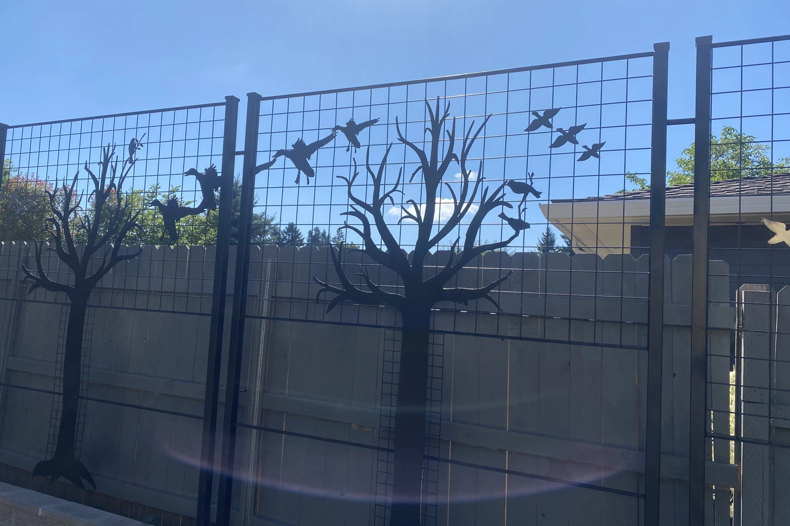 A black decorative metal art fence panel with a grid pattern featuring silhouette cutouts of bare trees and various birds in flight and perched on branches.