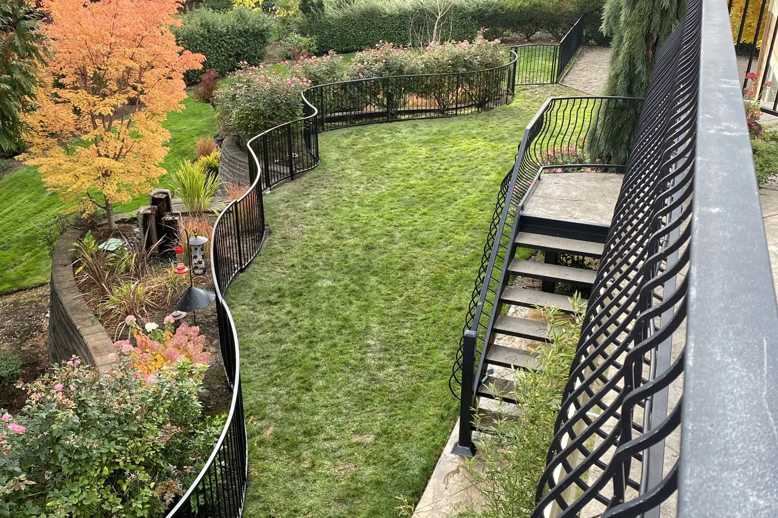 A custom-curved black ornamental iron fence bordering a lush green lawn and tiered stone retaining walls, alongside a black metal outdoor staircase. | Chip and Dale's Iron Works