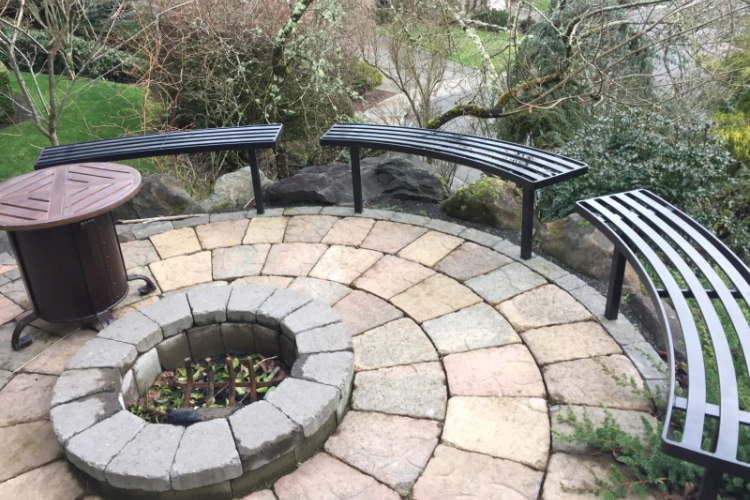 A set of curved black metal benches with a slat design, arranged around a circular stone paver patio and built-in fire pit in a residential backyard.