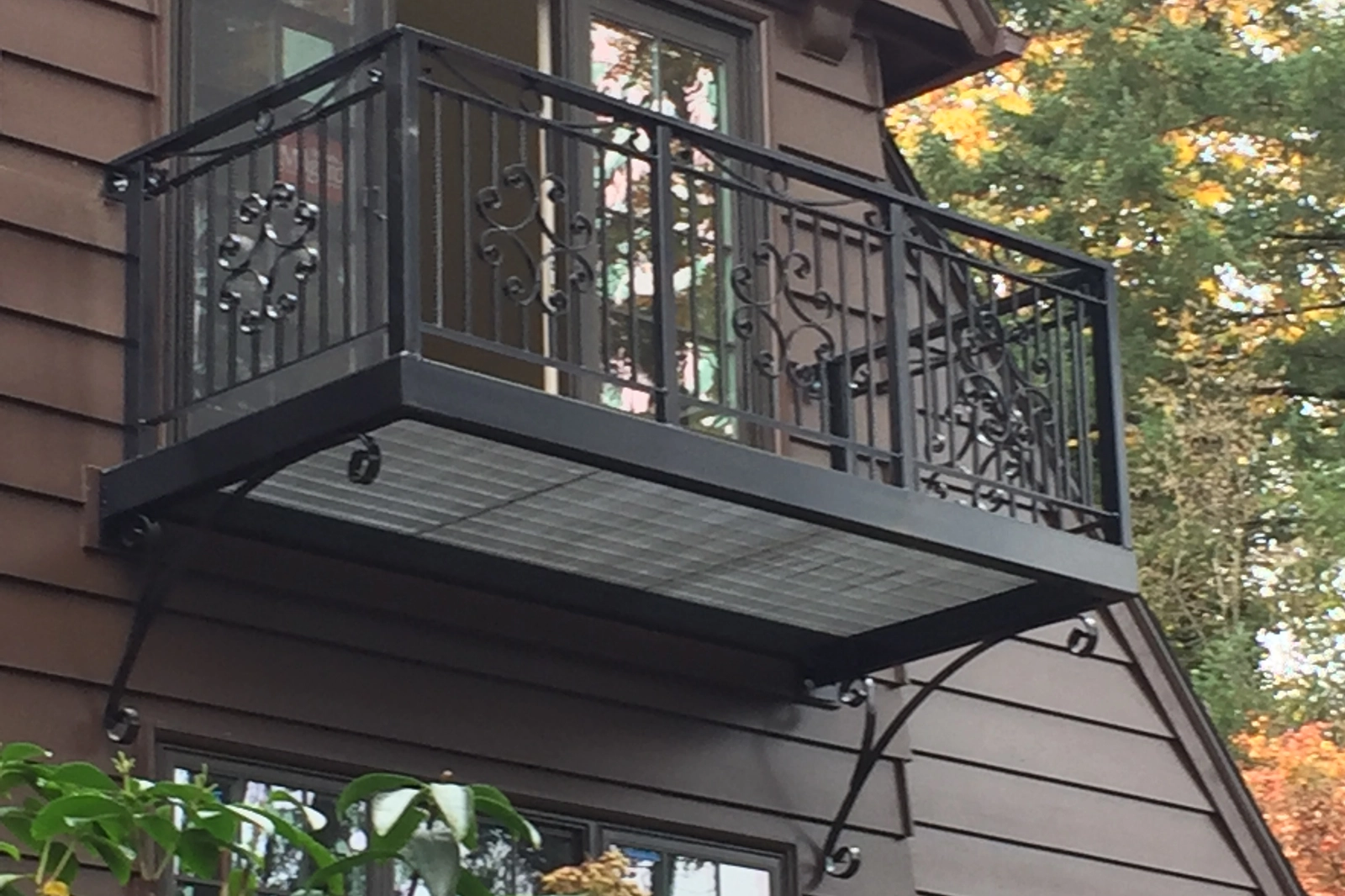 A black wrought iron Juliet balcony featuring decorative scrollwork panels, vertical pickets, and ornate support brackets on a residential home.
