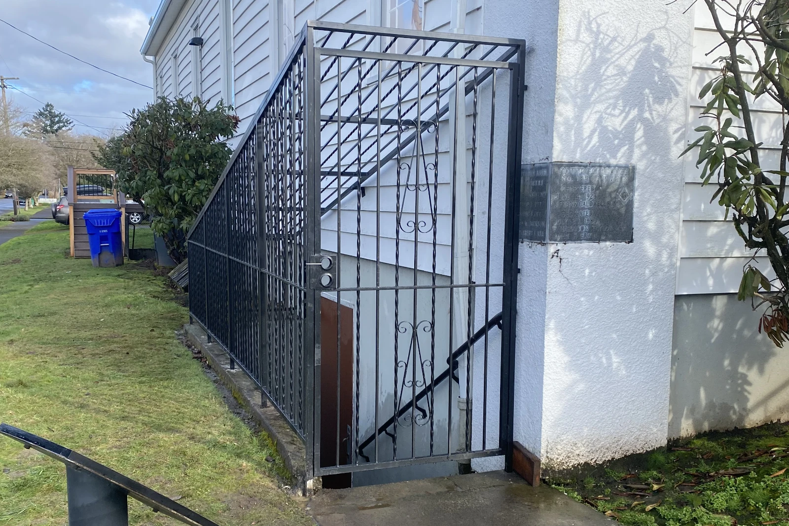 A custom-fabricated black wrought iron security enclosure with a tall pedestrian gate and twisted pickets, surrounding a concrete basement stairwell on a white residential building.