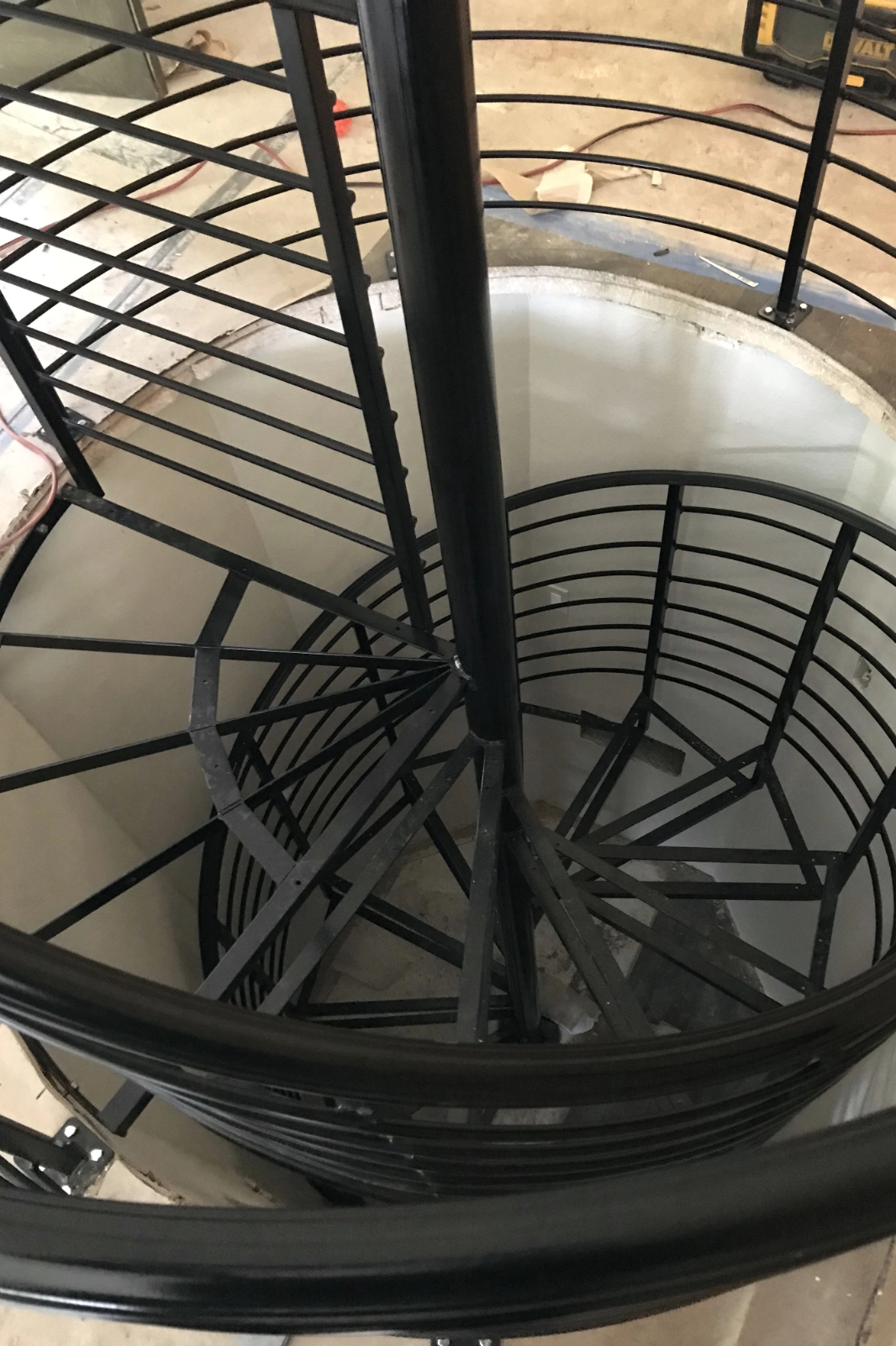 Top-down view of a custom-fabricated black metal spiral staircase being installed, featuring a central support pole and horizontal bar safety railings.