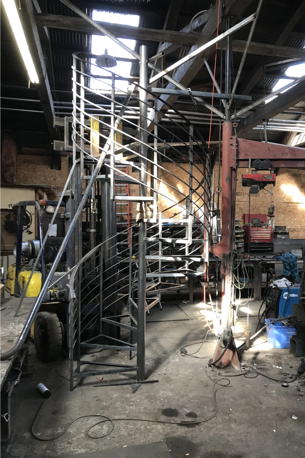A custom-fabricated raw steel spiral staircase being assembled in a metal shop, featuring a central support pole and sleek horizontal guardrail balusters.